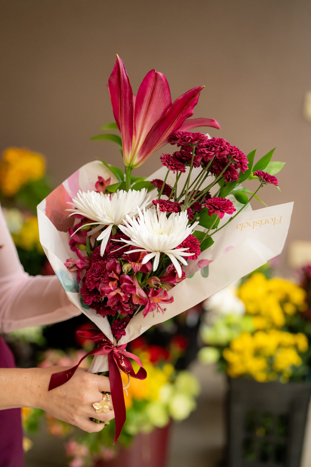 Buquê Florisbela com lírio magenta, crisântemos brancos, áster e alstromérias, embrulhado em papel artesanal e fita bordô.