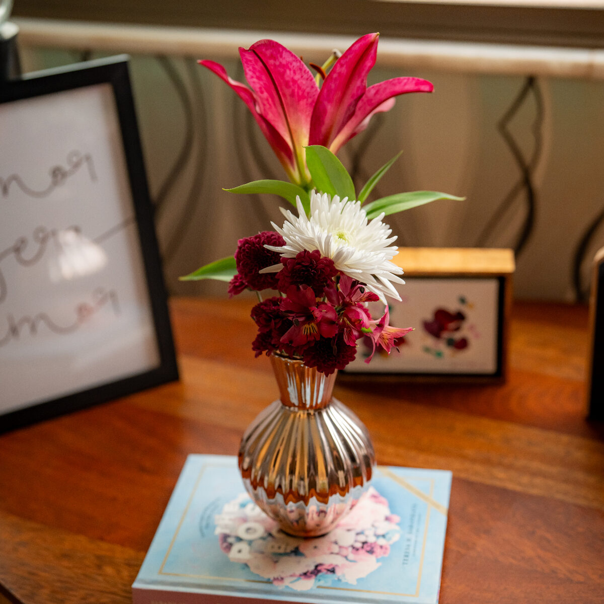 Arranjo Florisbela em vasinho metálico perolado com lírio magenta, crisântemo branco, mini crisântemos bordô e alstromérias, sobre o livro The Posy Book.