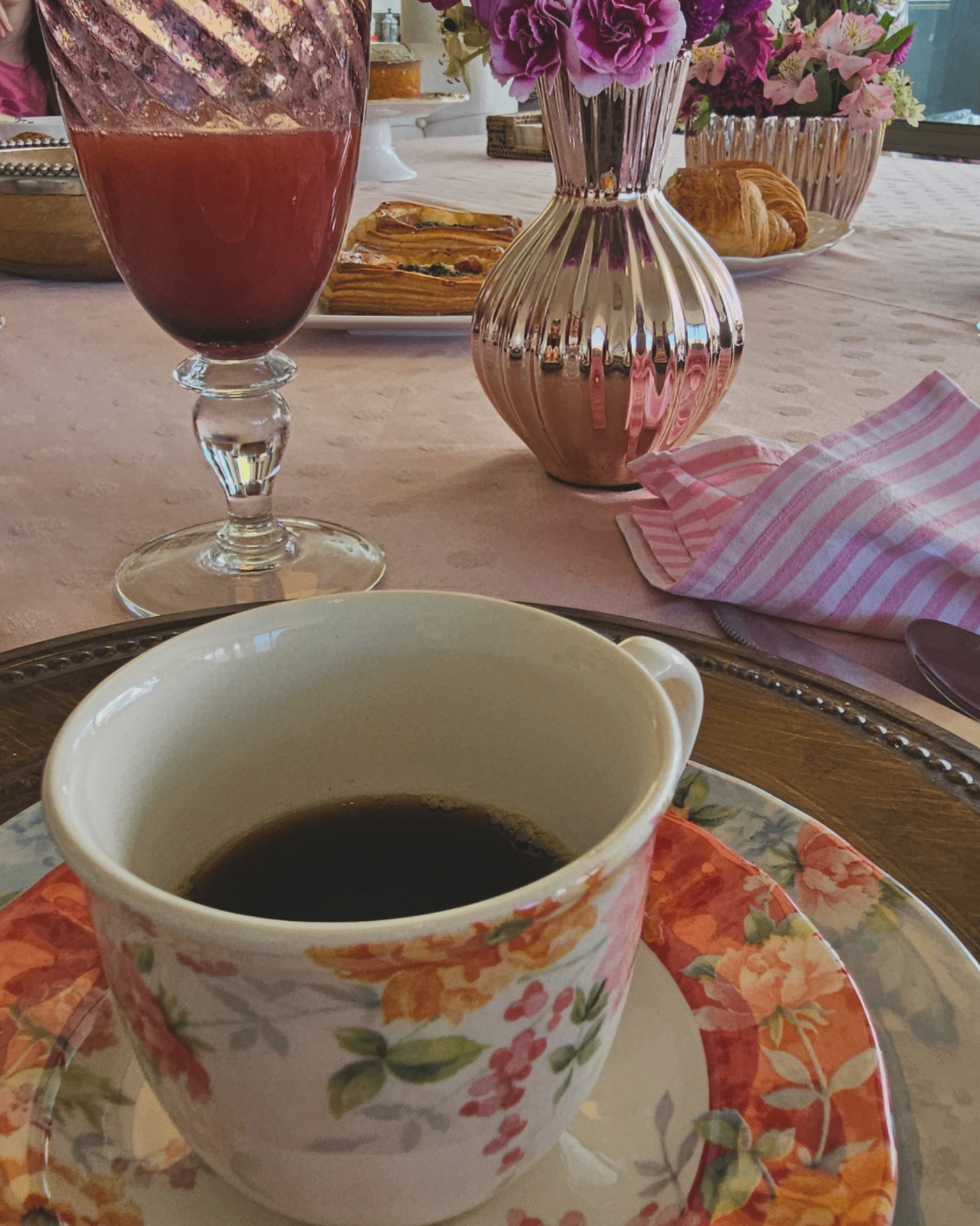 Xícara floral com café e arranjo Florisbela ao fundo.