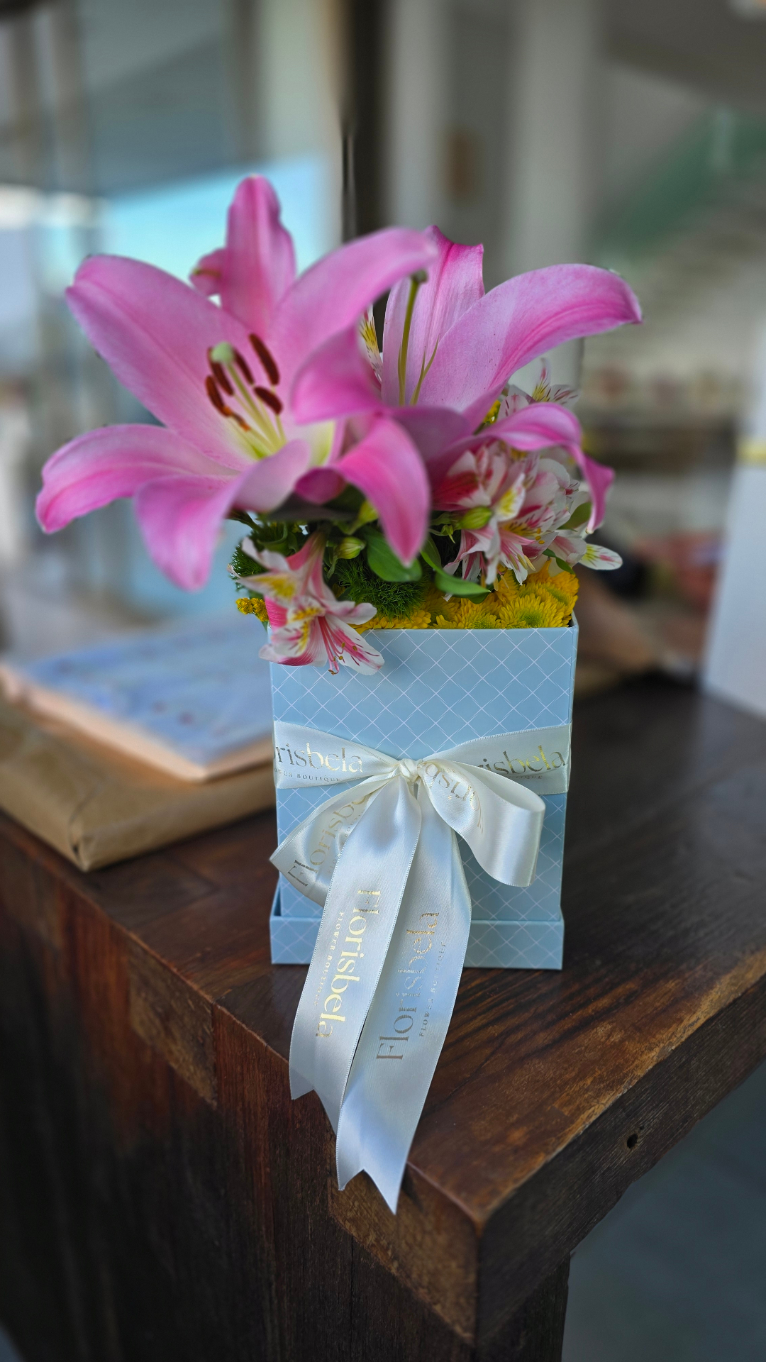 Arranjo em caixa azul Florisbela com lírios rosa, alstromérias e laço de fita branca.