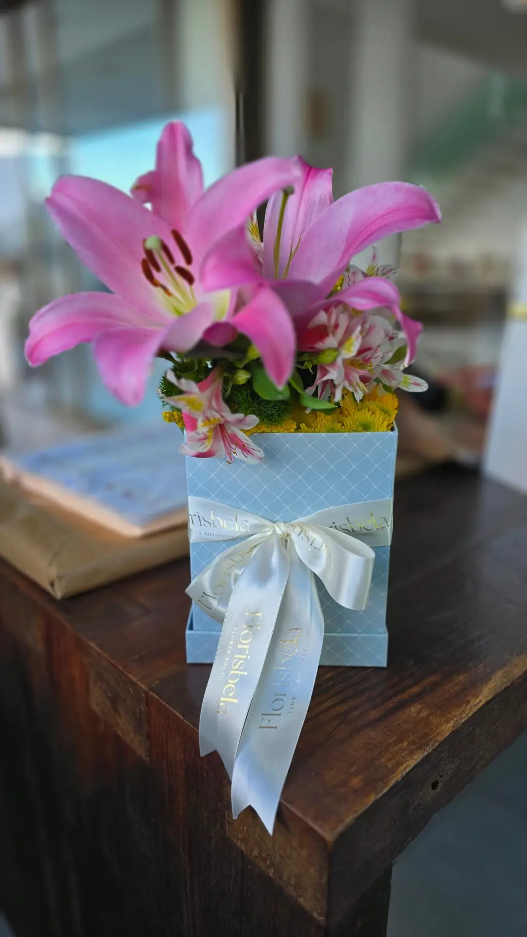 Arranjo em caixa azul Florisbela com lírios rosa, alstromérias e laço de fita branca.