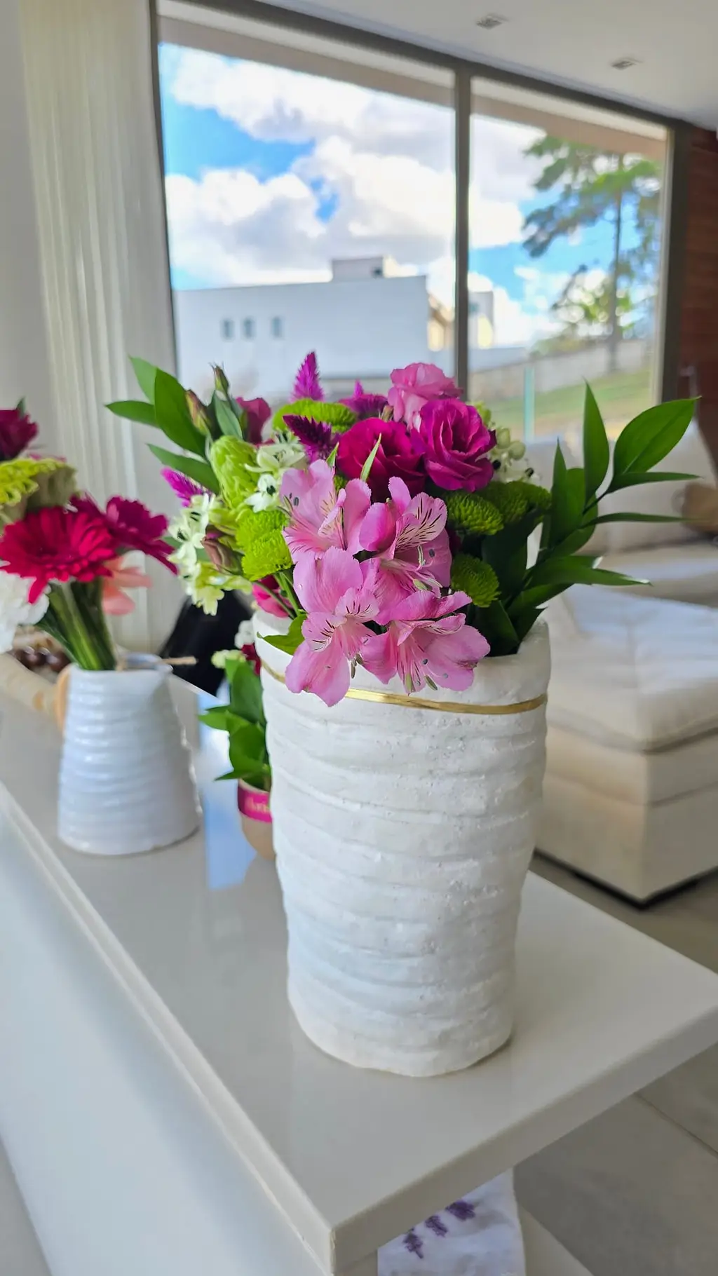 Arranjo de mesa em vaso branco artesanal com filete dourado e flores rosa.