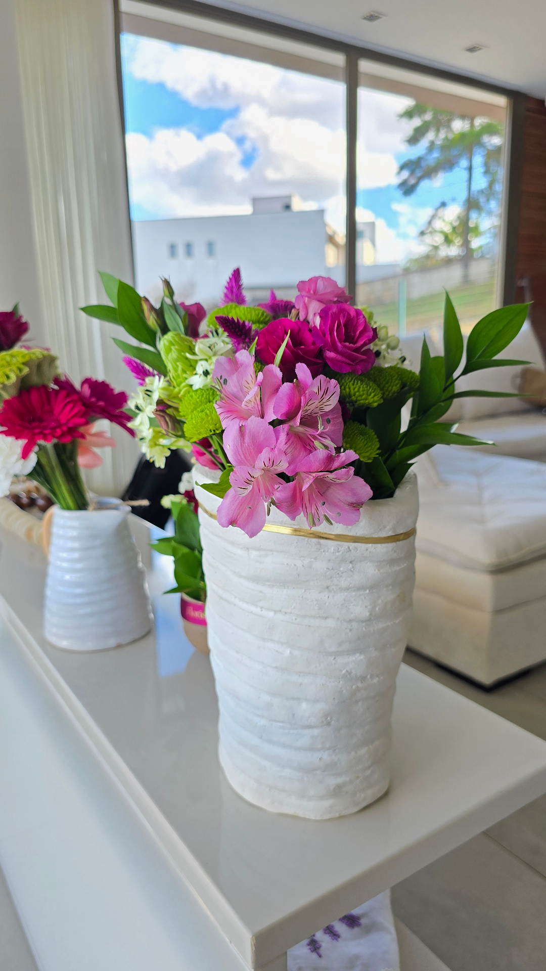 Arranjo de mesa em vaso branco artesanal com filete dourado e flores rosa.