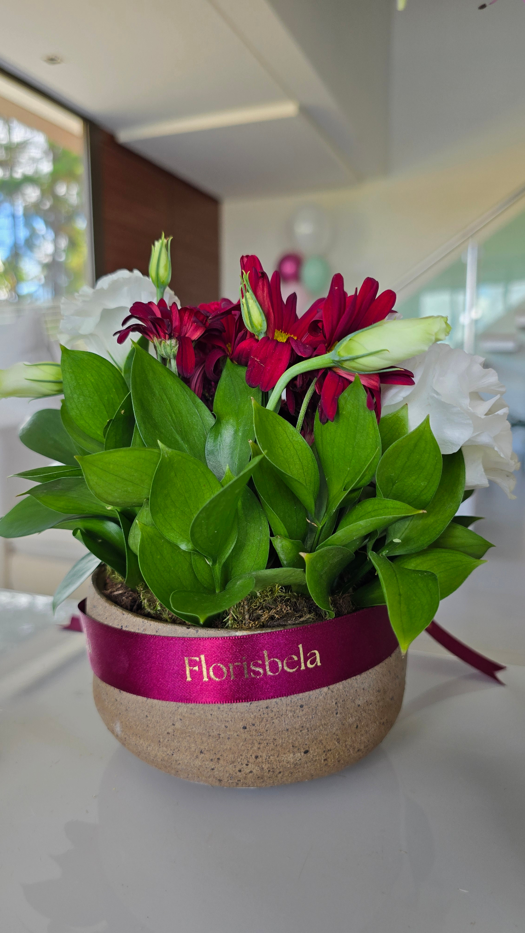 Arranjo de mesa em vaso de cerâmica com fita Florisbela, margaridas vermelhas e lisianthus brancos.