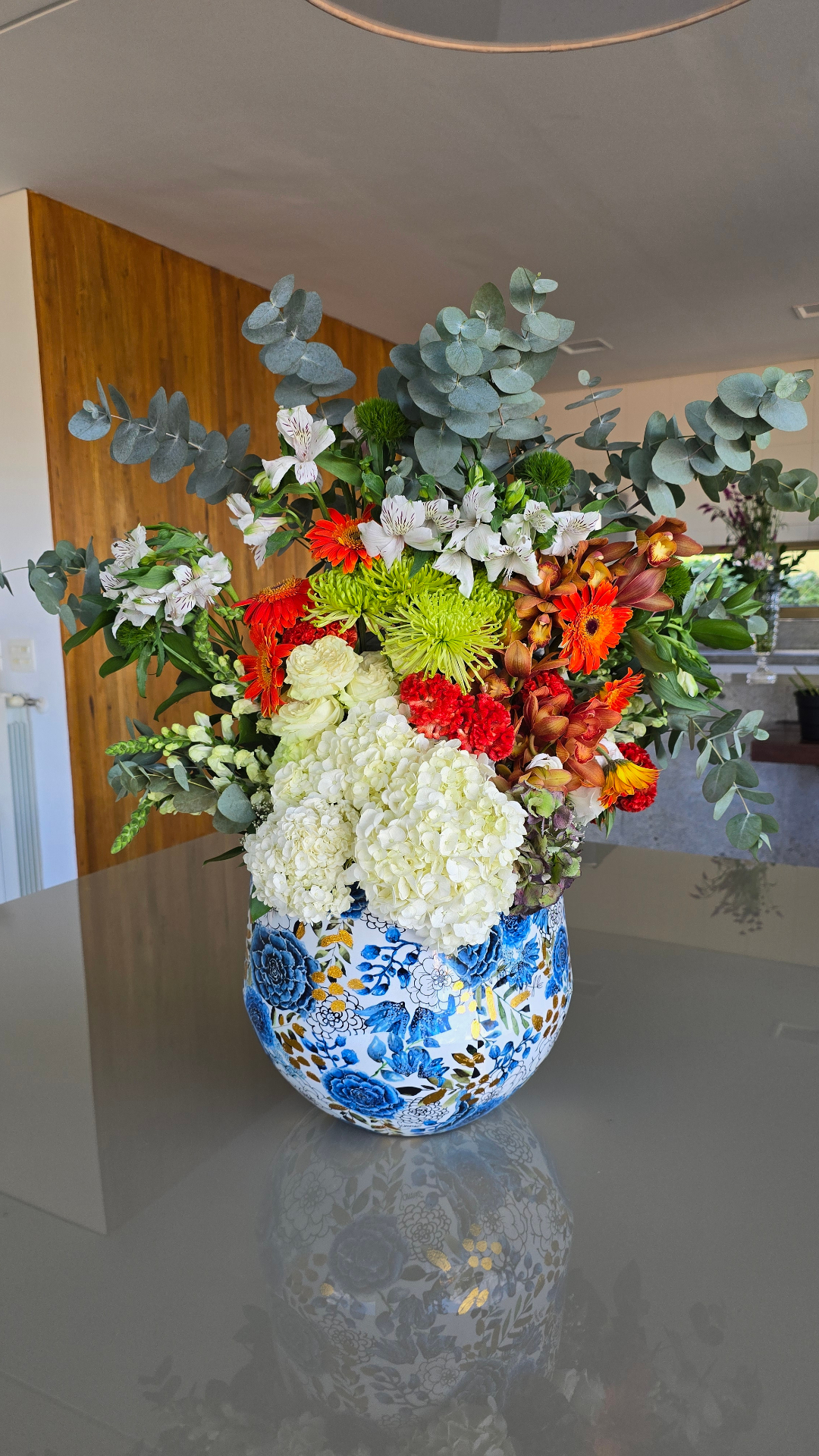 Arranjo exuberante em vaso azul floral com hortênsias brancas, gérberas laranja, crisântemo verde, orquídeas cymbidium e eucalipto.